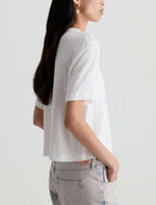 A woman shown from the side wearing a white short-sleeve cropped t-shirt and light-wash jeans against a plain light gray background.