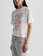 A woman wearing a white cropped t-shirt with a colorful graphic print and light-wash jeans, photographed against a plain light gray studio background.