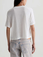 A woman viewed from the back wearing a short-sleeve white knit crop top and light-wash jeans against a plain light gray studio background.