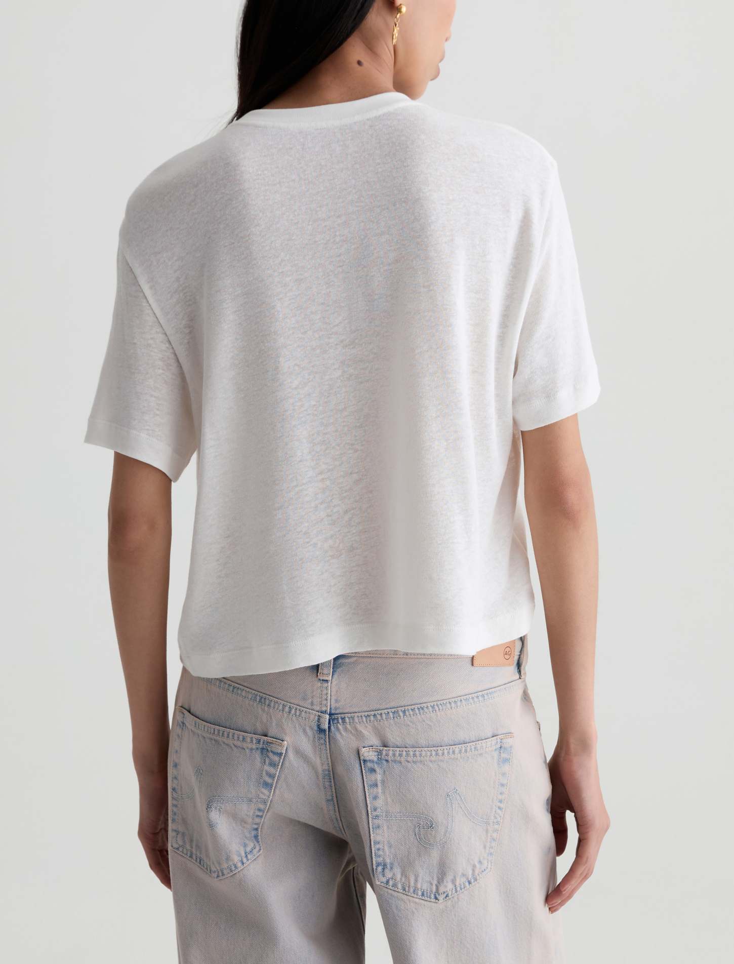 A woman viewed from the back wearing a short-sleeve white knit crop top and light-wash jeans against a plain light gray studio background.
