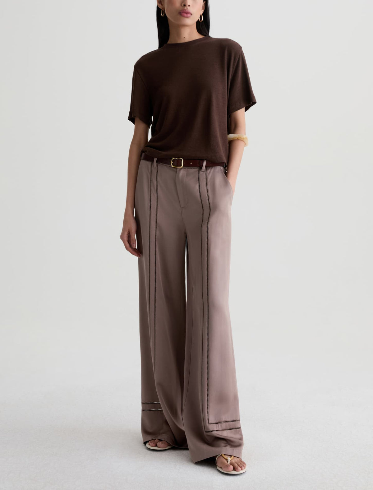 A model wearing a dark brown short-sleeve t-shirt and wide-leg taupe trousers, styled with a brown belt and flat sandals against a light gray studio background.