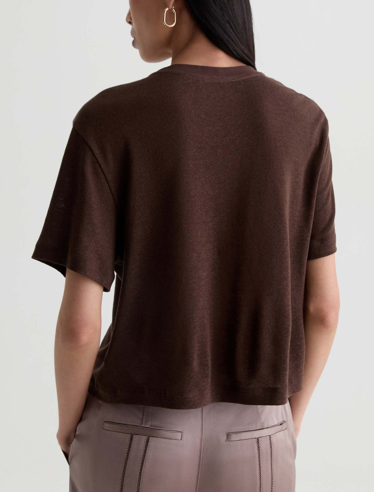 A woman shown from the back wearing a dark brown short-sleeve knit top and light gray trousers against a plain light gray background.