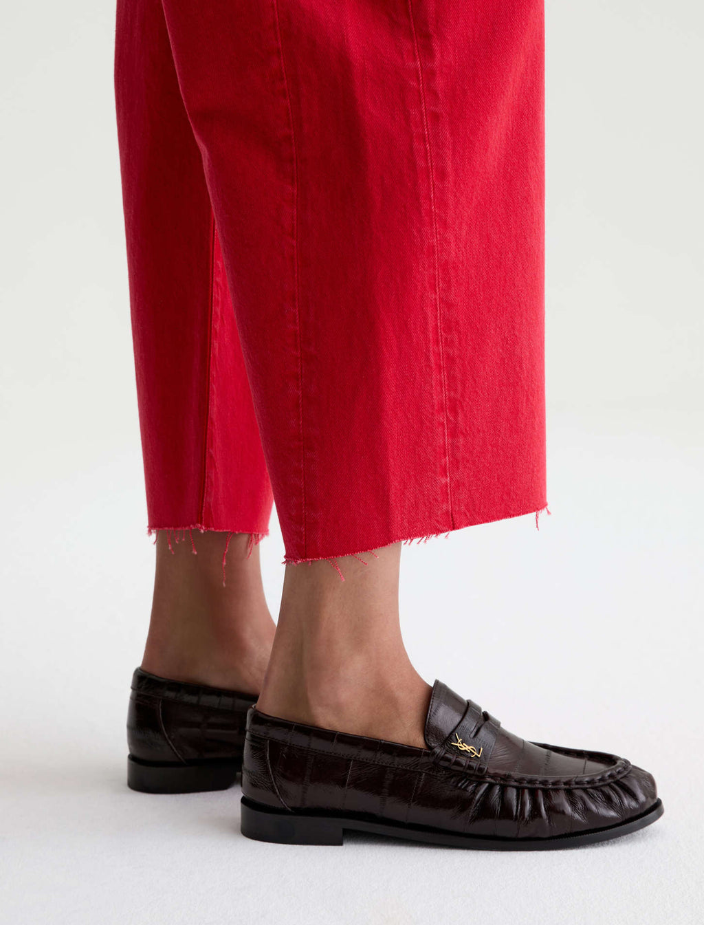 A close-up of a person's feet wearing dark brown crocodile-embossed leather penny loafers and bright red cropped jeans against a clean white background.