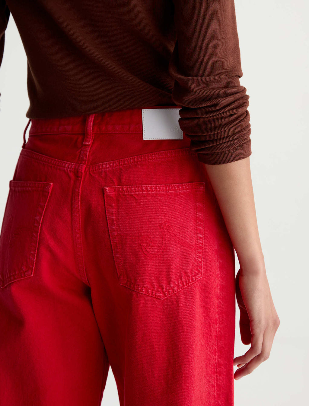 Close-up back view of a person wearing bright red jeans and a brown ribbed long-sleeve top against a light gray studio background.