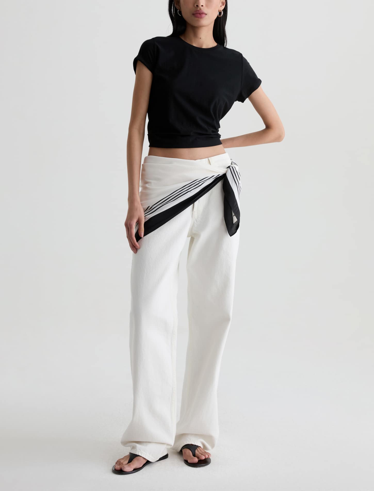 A model wearing a black cropped short-sleeve t-shirt and white wide-leg trousers with a black and white striped scarf tied around the waist, paired with black sandals, against a light gray studio background.