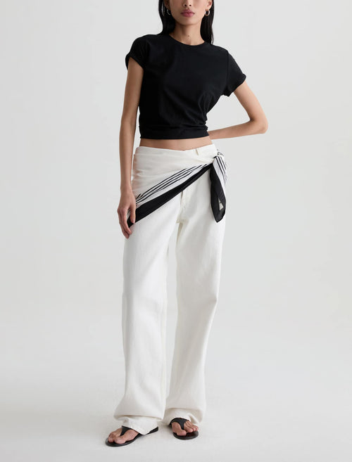 A model wearing a black cropped short-sleeve t-shirt and white wide-leg trousers with a black and white striped scarf tied around the waist, paired with black sandals, against a light gray studio background.