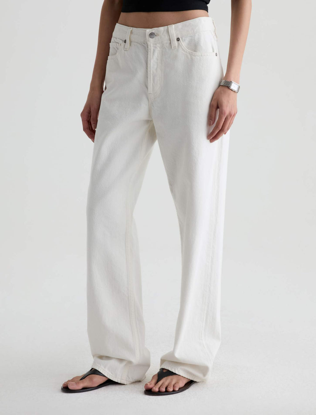 A model wearing a pair of off-white, wide-leg denim jeans with a black crop top and black flip-flops, shown from the waist down against a light gray studio background.