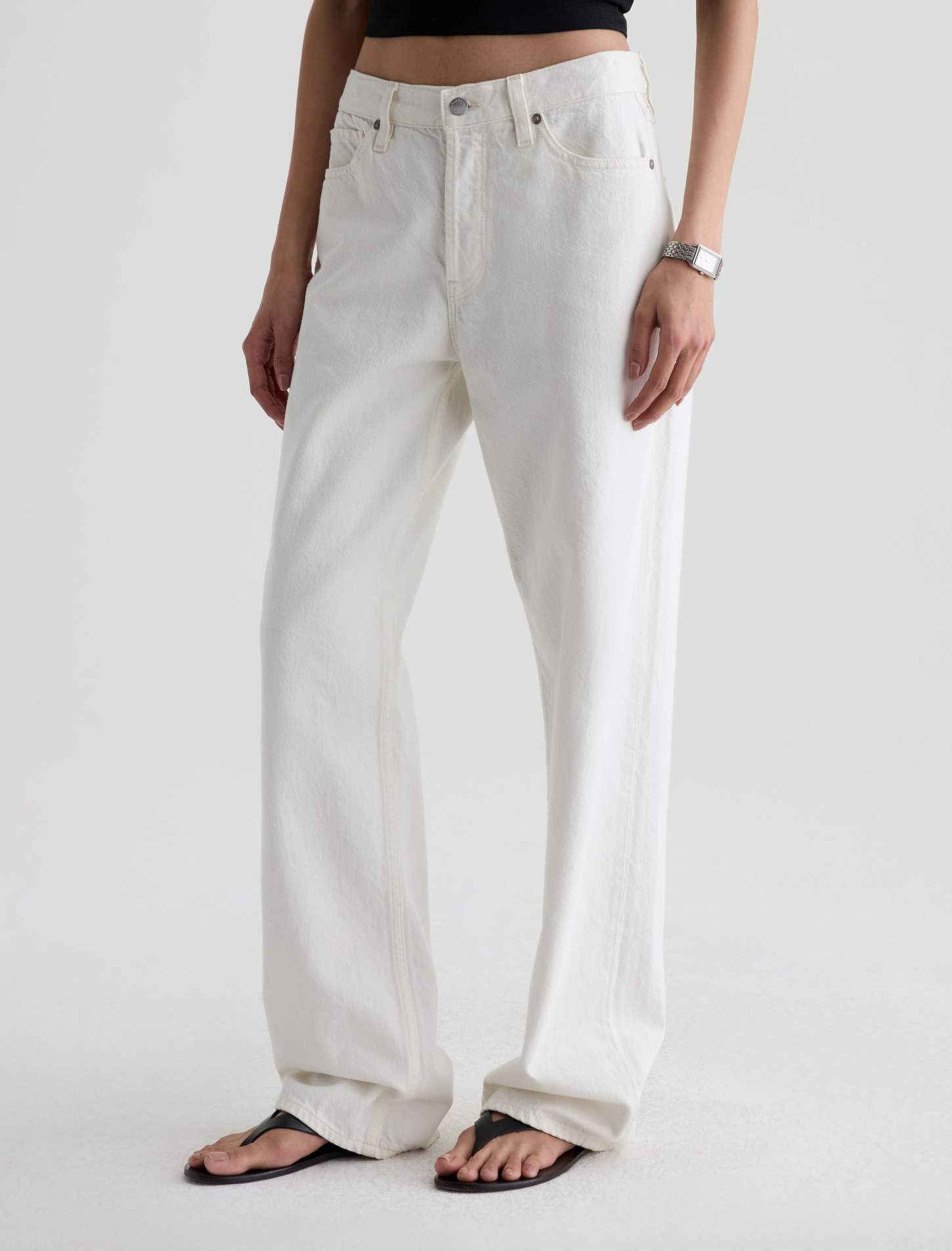 A model wearing a pair of off-white, wide-leg denim jeans with a black crop top and black flip-flops, shown from the waist down against a light gray studio background.