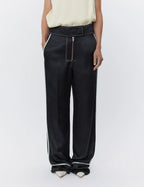 A model wearing black satin pants with white piping trim and a burnt red metal zip at the center front.