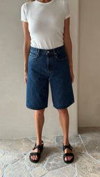 A pair of blue denim shorts with a button-up front, worn by a person standing indoors.