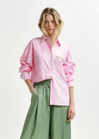 A woman wearing an oversized pink shirt with vertical white stripes, featuring a patch pocket with rhinestone embellishments, paired with green trousers.