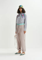 A model wearing organic cotton floral-print pants with a double-waisted design and contrasting elasticated waistband and drawstring fastening, along with a grey shirt with colorful embroidery and a hat.
