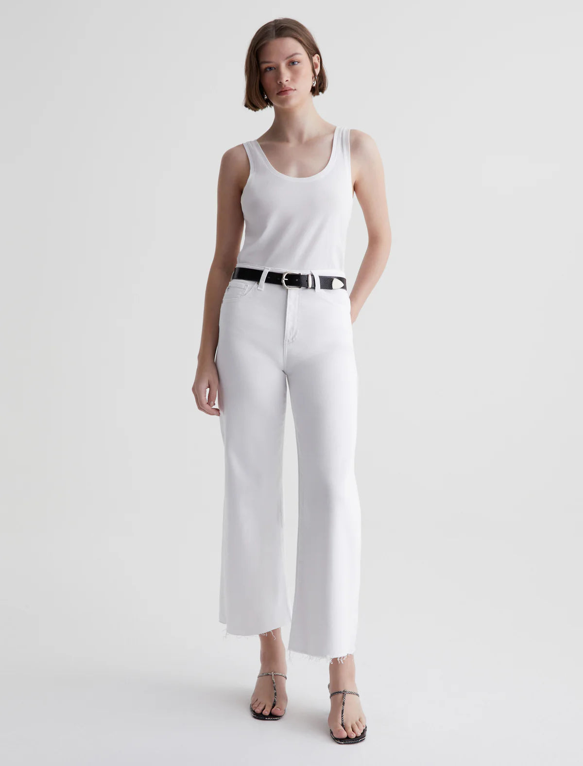 A person standing wearing mid-rise white wide leg crop jeans and a sleeveless top, accessorized with a belt and earrings.
