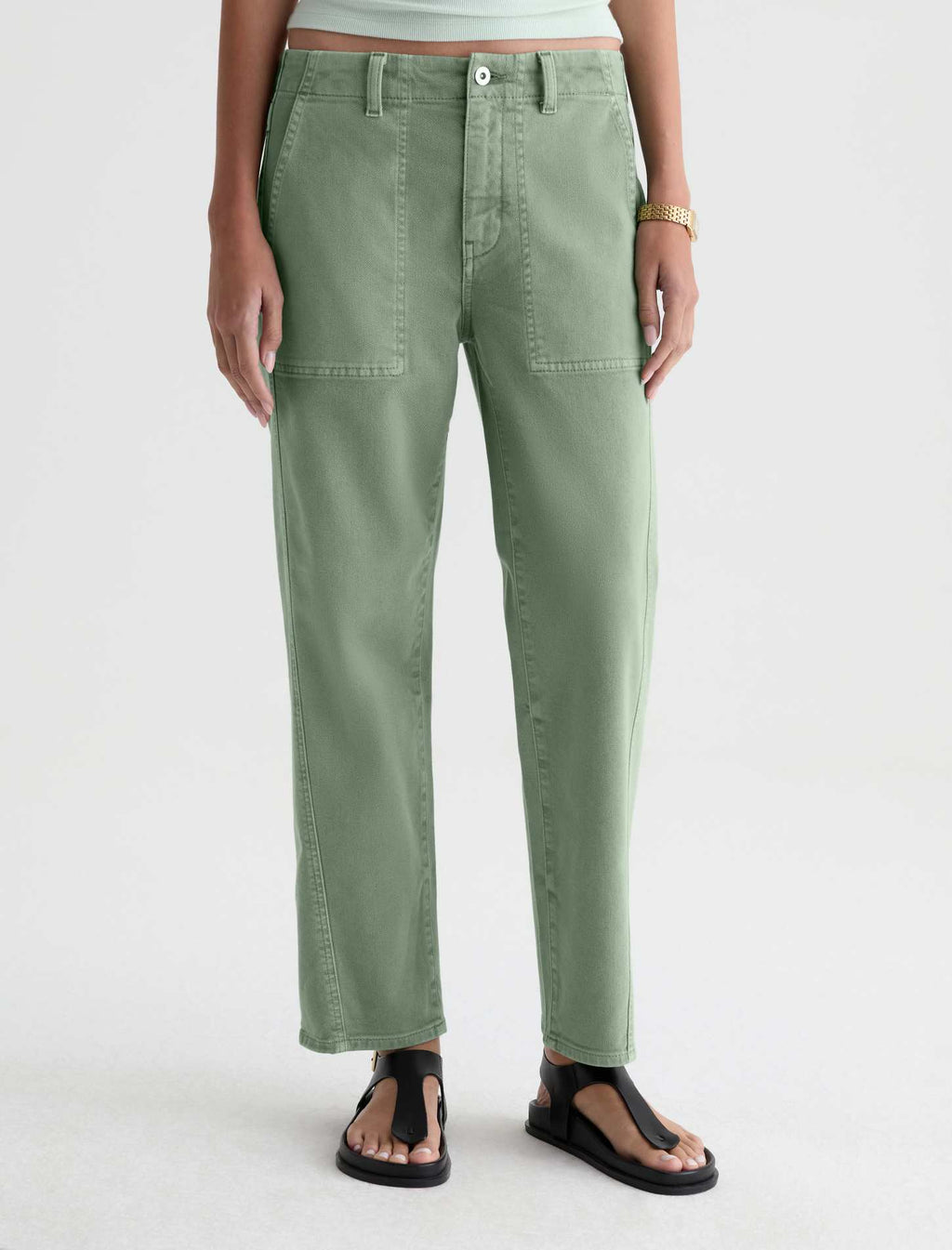 A person wearing sage green straight-leg pants with front patch pockets, shown from the waist down against a clean light gray studio background. The person is also wearing black thong sandals and a light top, but the product focus appears to be the pants.