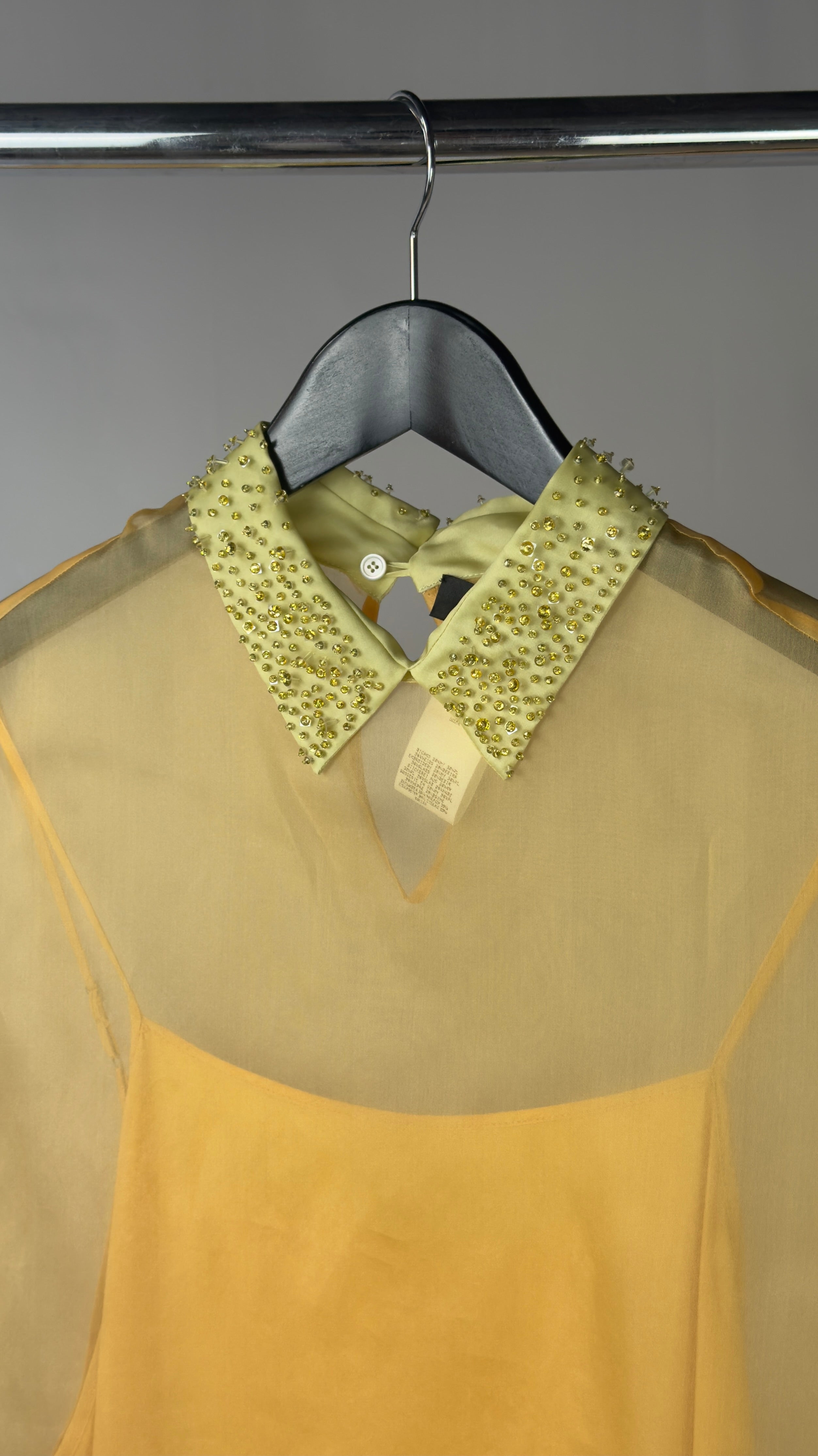 Sheer beige blouse with green sequin collar on a hanger against a gray background