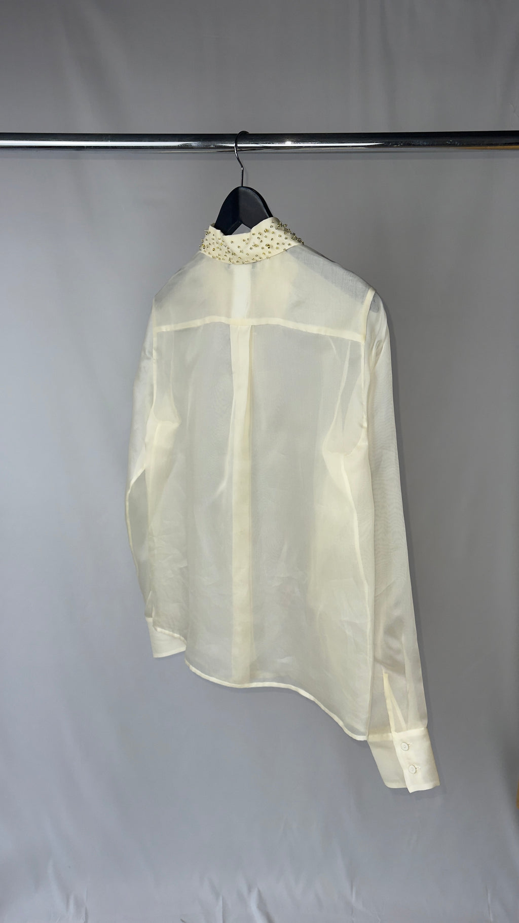 Sheer white blouse with embellishments on a hanger against a gray background
