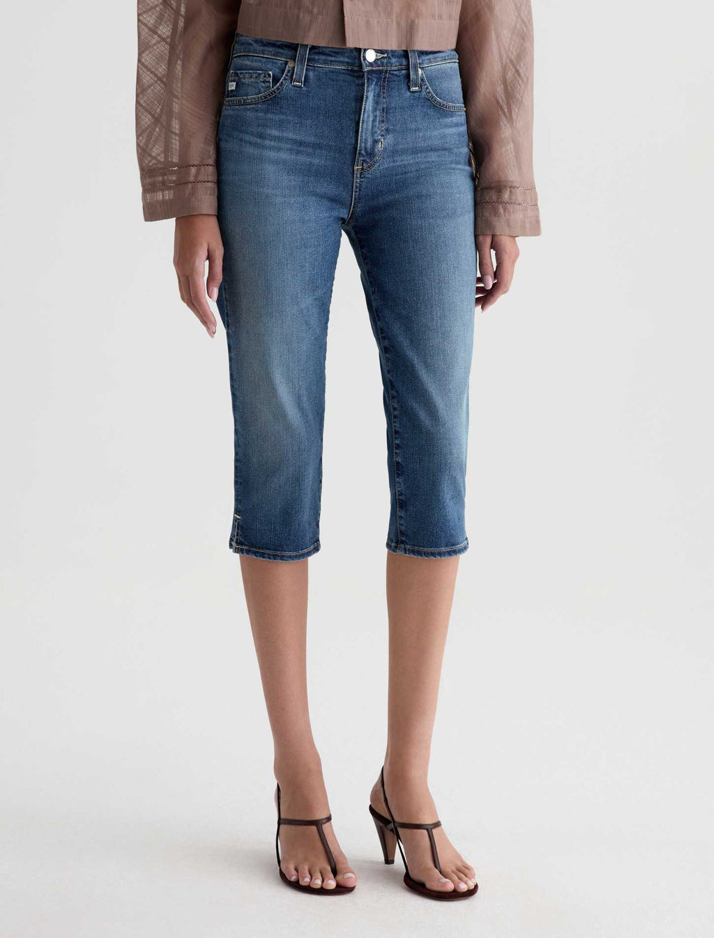 A model is shown from the waist down wearing medium-wash blue cropped denim jeans, a brown patterned blouse, and black strappy heels against a light gray studio background.