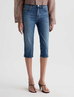 A model is shown from the waist down wearing medium-wash blue cropped denim jeans, a brown patterned blouse, and black strappy heels against a light gray studio background.