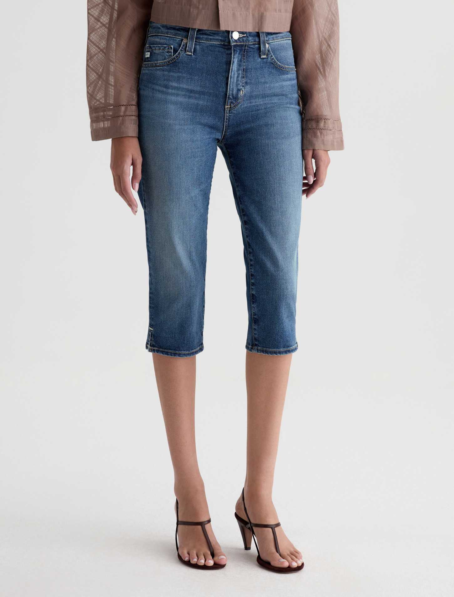 A model is shown from the waist down wearing medium-wash blue cropped denim jeans, a brown patterned blouse, and black strappy heels against a light gray studio background.