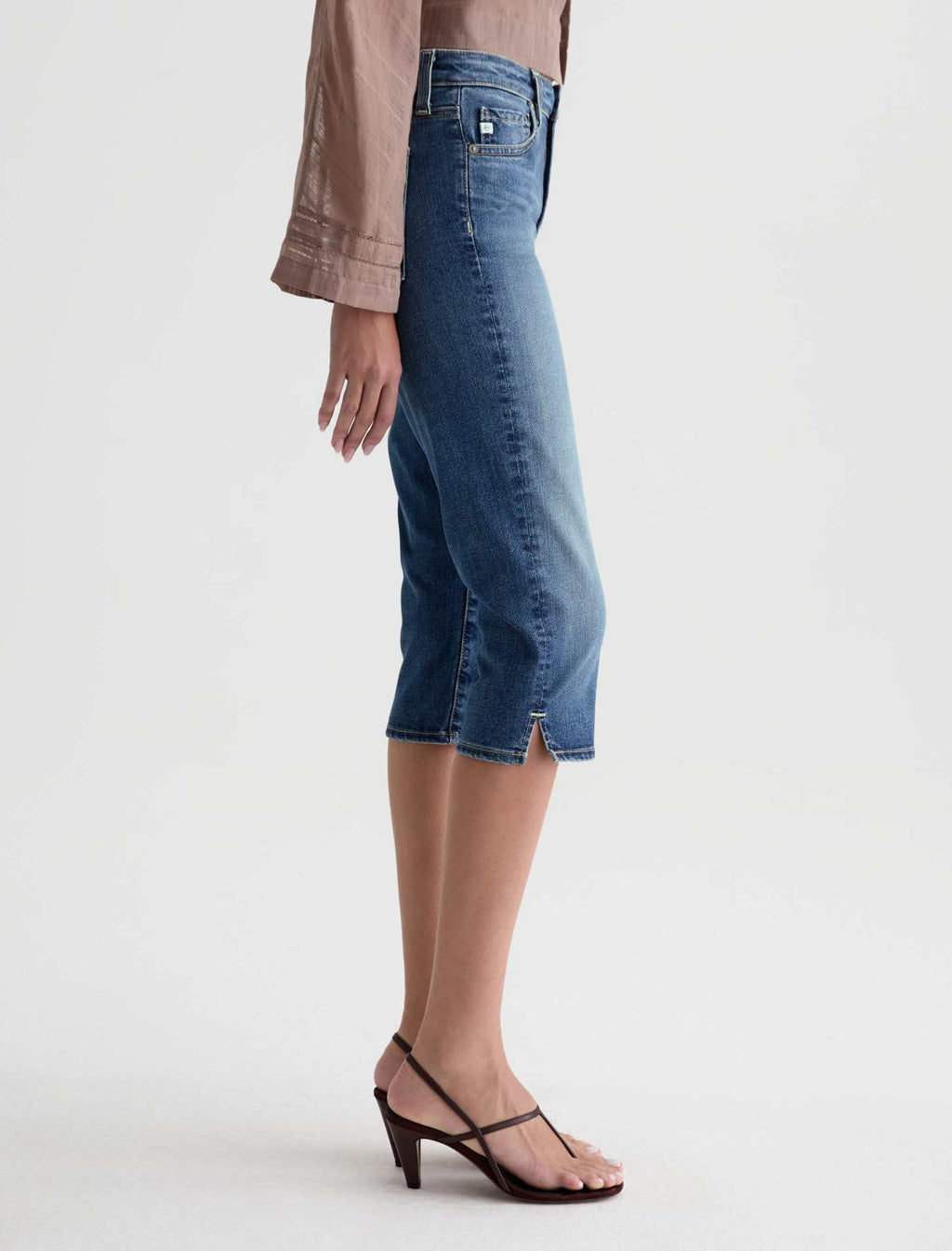 A side-profile view of a model wearing medium-wash blue denim capri pants with a side slit, styled with a taupe long-sleeve blouse and dark brown strappy sandals against a light gray studio background.