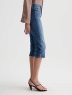 A side-profile view of a model wearing medium-wash blue denim capri pants with a side slit, styled with a taupe long-sleeve blouse and dark brown strappy sandals against a light gray studio background.