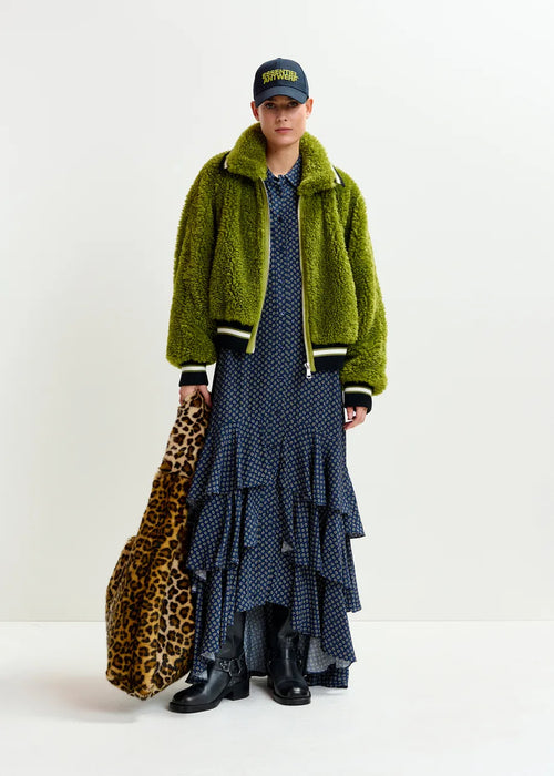 Person wearing a green jacket, blue polka dot dress, and black boots holding a leopard print bag on a white background