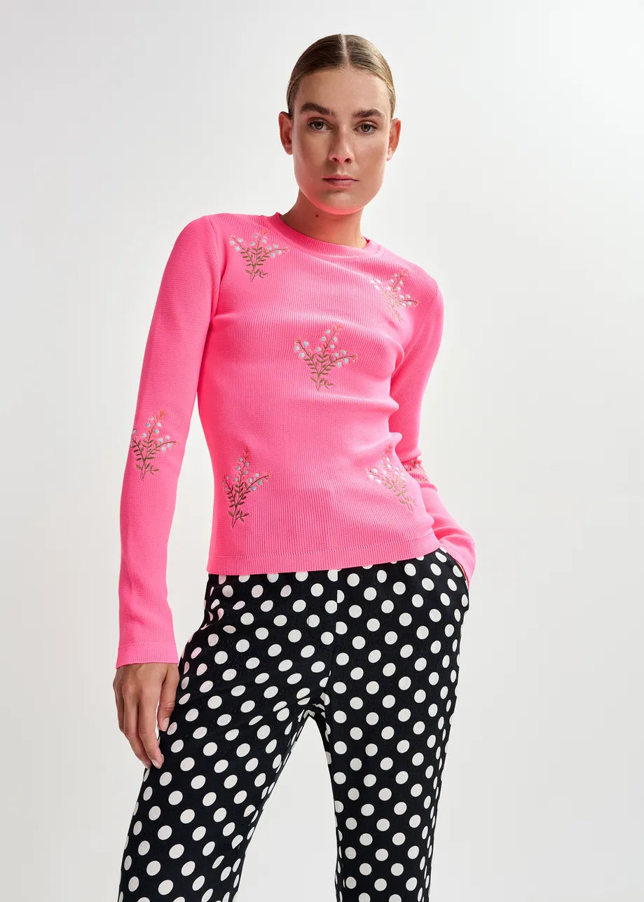 Person wearing a pink long-sleeve top with floral embellishments and black polka dot pants on a light gray background.JARDENS-CO17