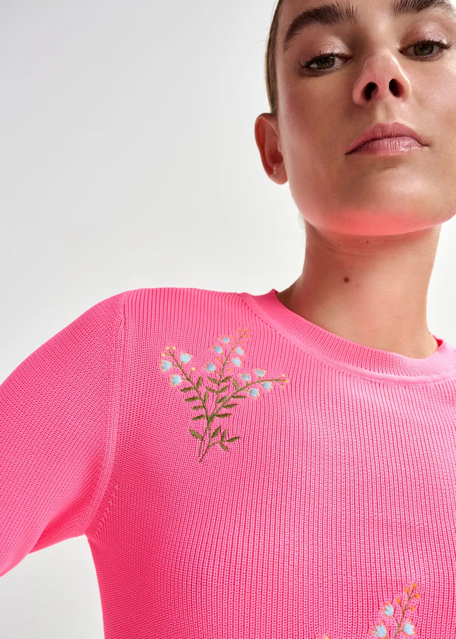 Person wearing a pink sweater with floral embroidery on a plain background