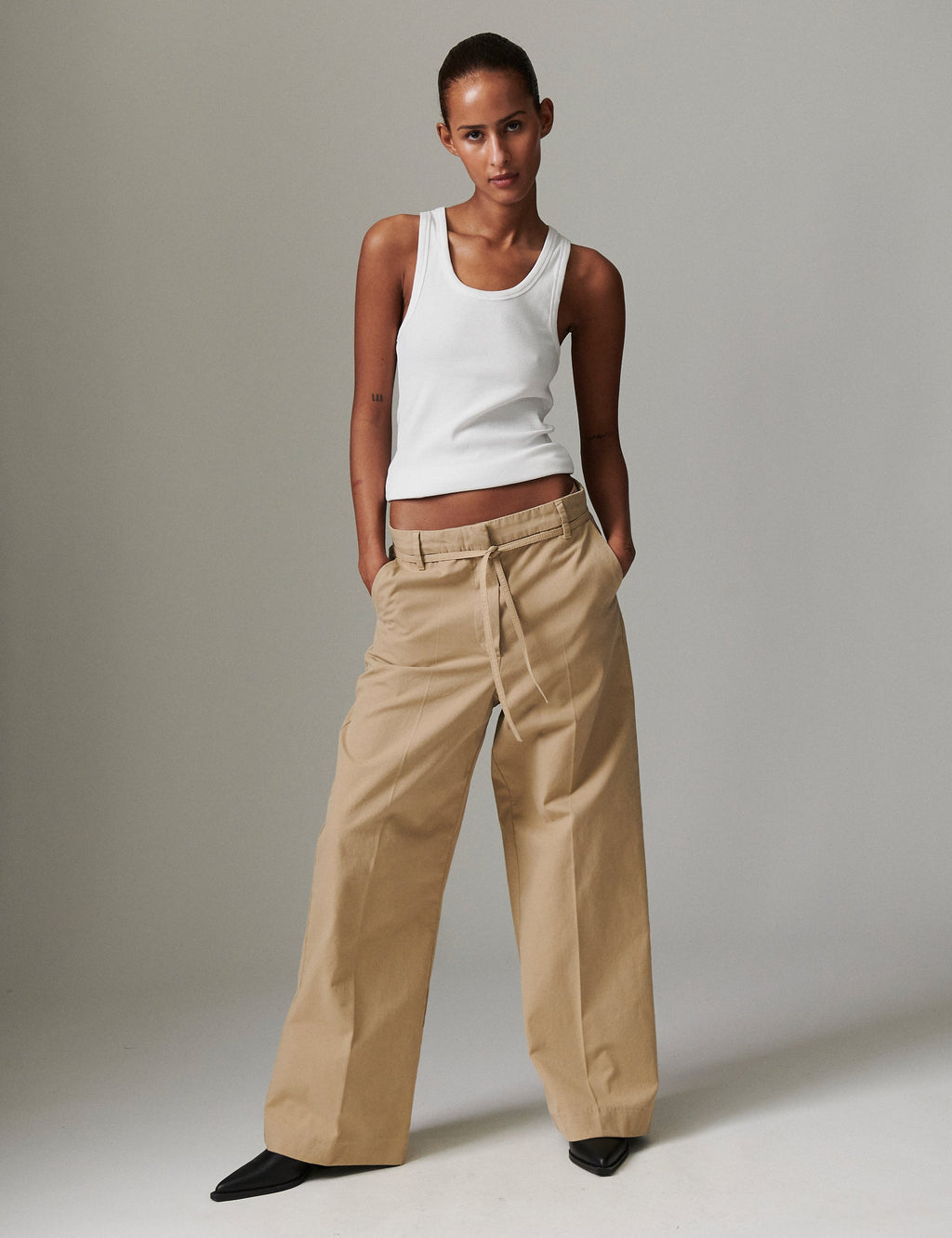 Woman wearing a white tank top and beige pants against a plain background