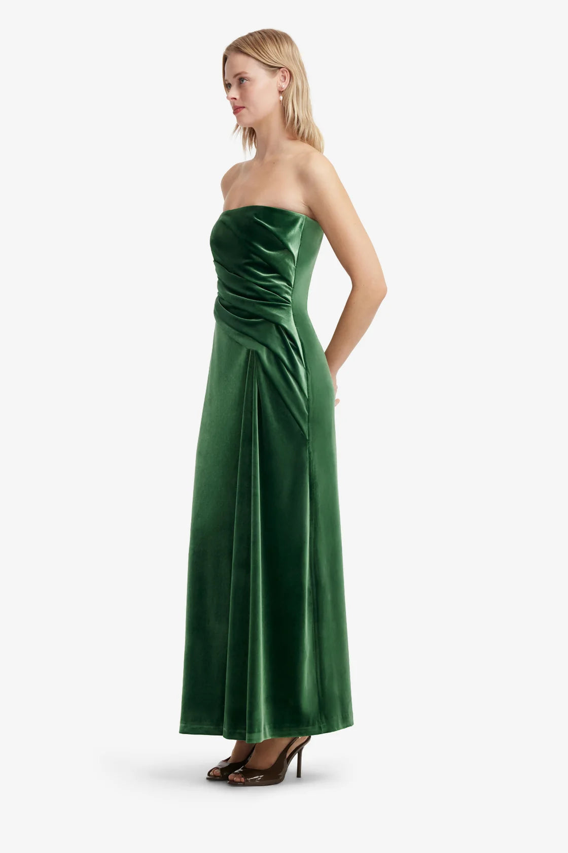 A woman modeling a strapless emerald green velvet maxi dress with a ruched bodice and draped skirt, shown against a plain light gray studio background.