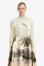 Woman wearing a light-colored blouse with a tie collar and a scenic skirt on a white background