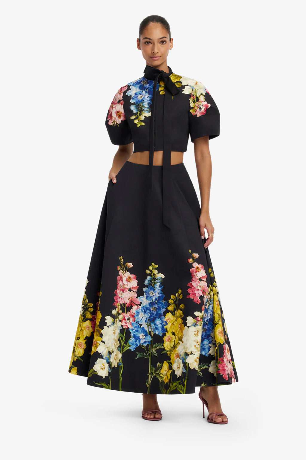A model wearing a black two-piece outfit featuring a short puff-sleeve crop top with a colorful floral print and a matching high-waisted flared skirt with a tie waist and a large floral border print.