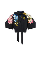 A black short-sleeve button-front blouse with a large bow at the collar, a tie waist, and colorful floral prints on the chest and both sleeves, shown on a plain light gray background.