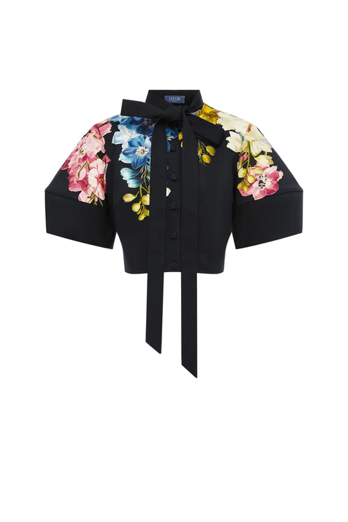 A black short-sleeve button-front blouse with a large bow at the collar, a tie waist, and colorful floral prints on the chest and both sleeves, shown on a plain light gray background.