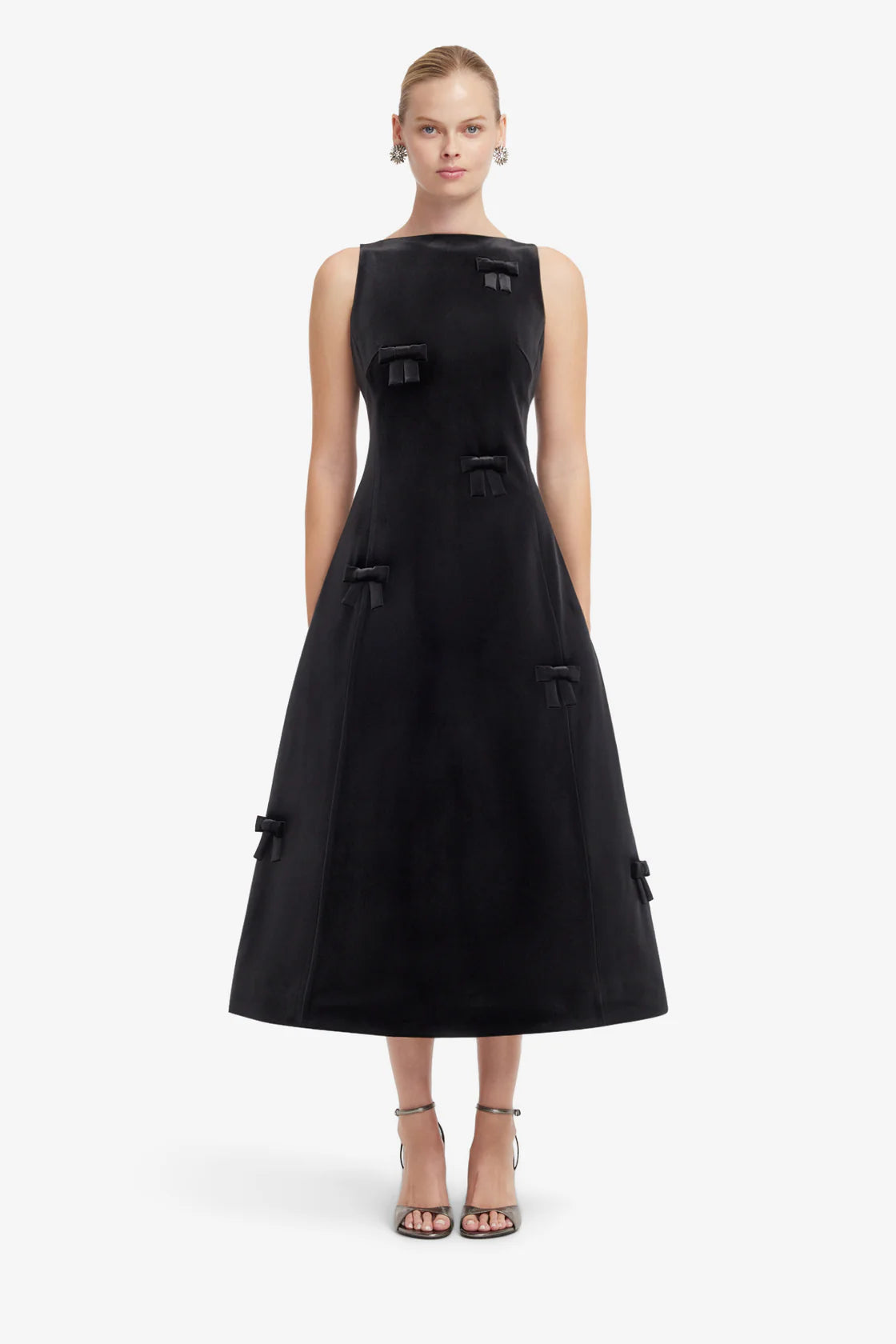A model wearing a sleeveless black midi dress with structured shoulders, decorative bows, and a flared skirt, paired with strappy high-heeled sandals against a light gray studio background.