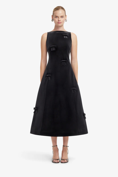 A model wearing a sleeveless black midi dress with structured shoulders, decorative bows, and a flared skirt, paired with strappy high-heeled sandals against a light gray studio background.