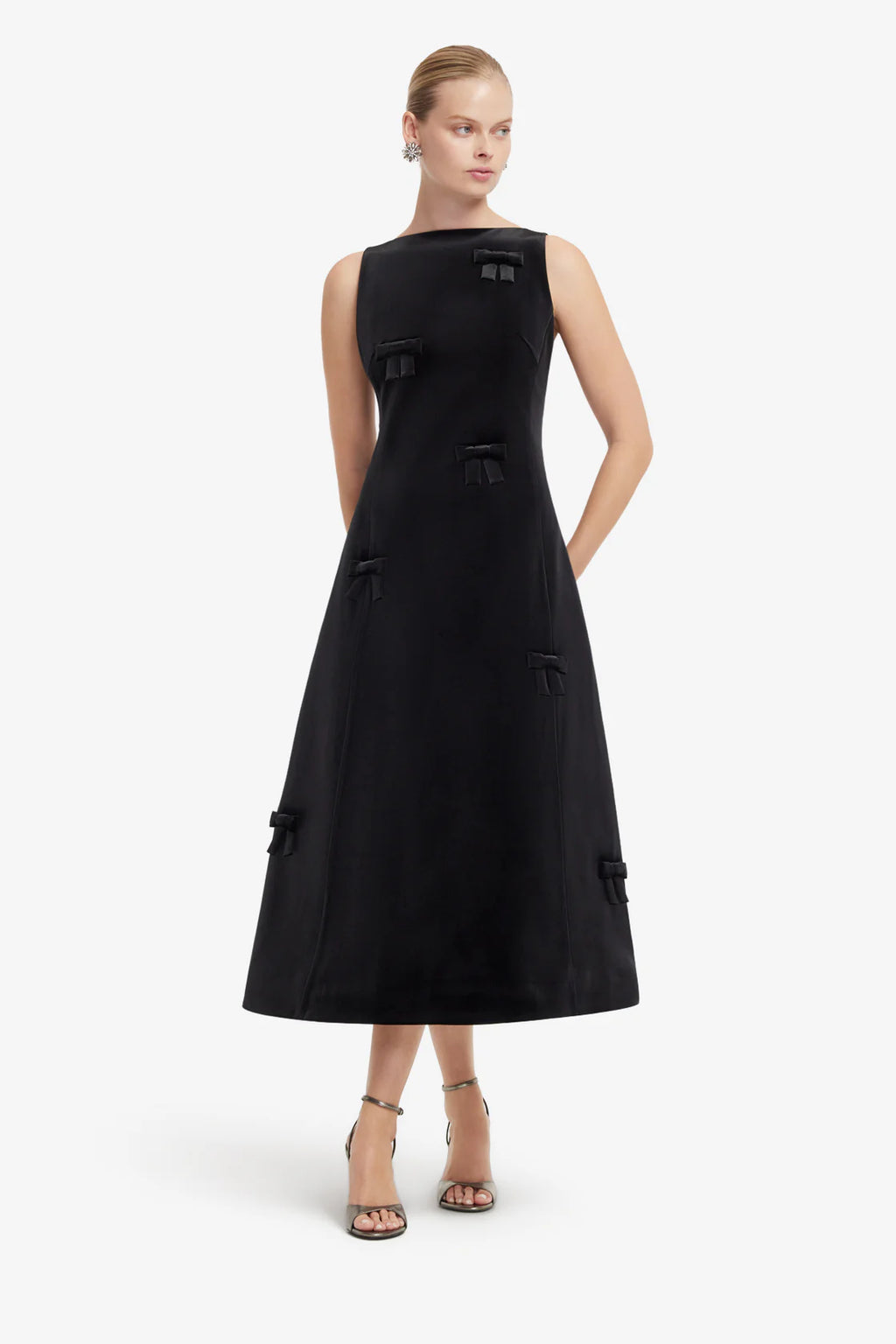 A woman modeling a sleeveless black midi dress with structured shoulders, decorative bow details, and side pockets against a plain light gray studio background.