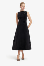 A woman modeling a sleeveless black midi dress with structured shoulders, decorative bow details, and side pockets against a plain light gray studio background.