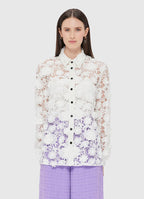 A woman wearing a white and purple floral lace shirt with a collared neckline and slim sleeves, with a front button opening. She is also wearing purple trousers.