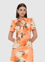 A woman wearing a short-sleeved, collared, tangerine orange mini dress with a floral hydrangea print, featuring front button closure.