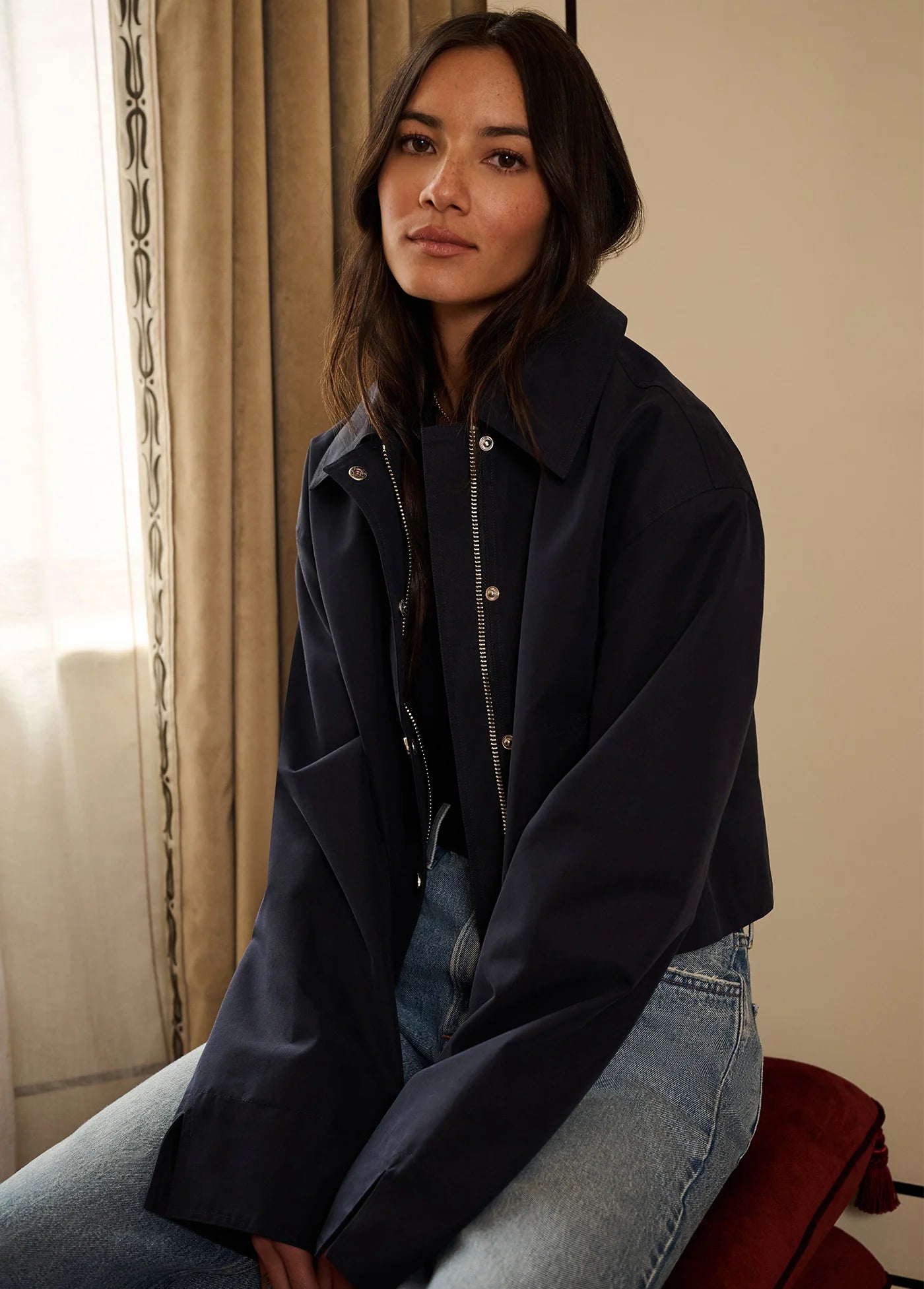 A woman wearing a dark navy oversized jacket over a black top and light blue jeans, seated indoors near a window with beige curtains.