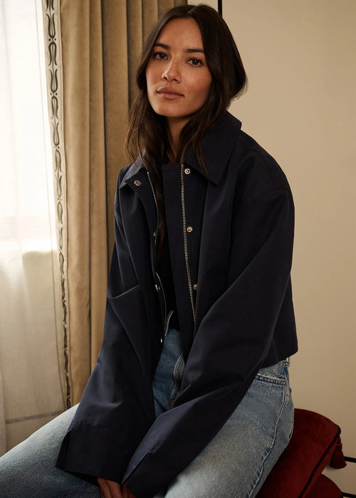 A woman wearing a dark navy oversized jacket over a black top and light blue jeans, seated indoors near a window with beige curtains.
