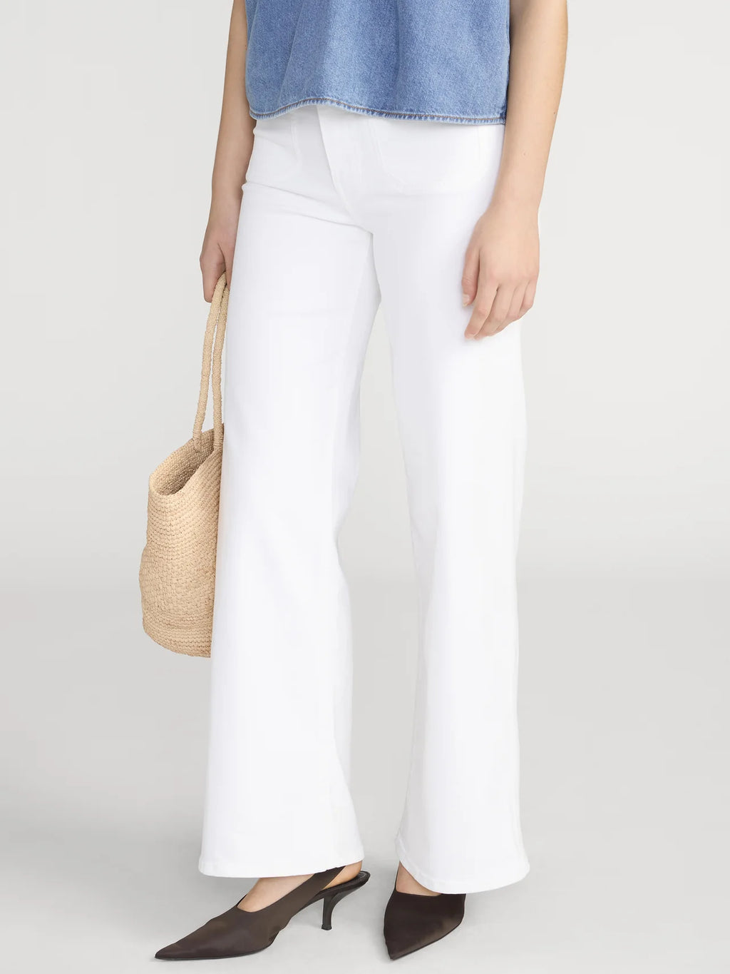Person wearing white pants and a denim top, holding a woven bag on a plain background