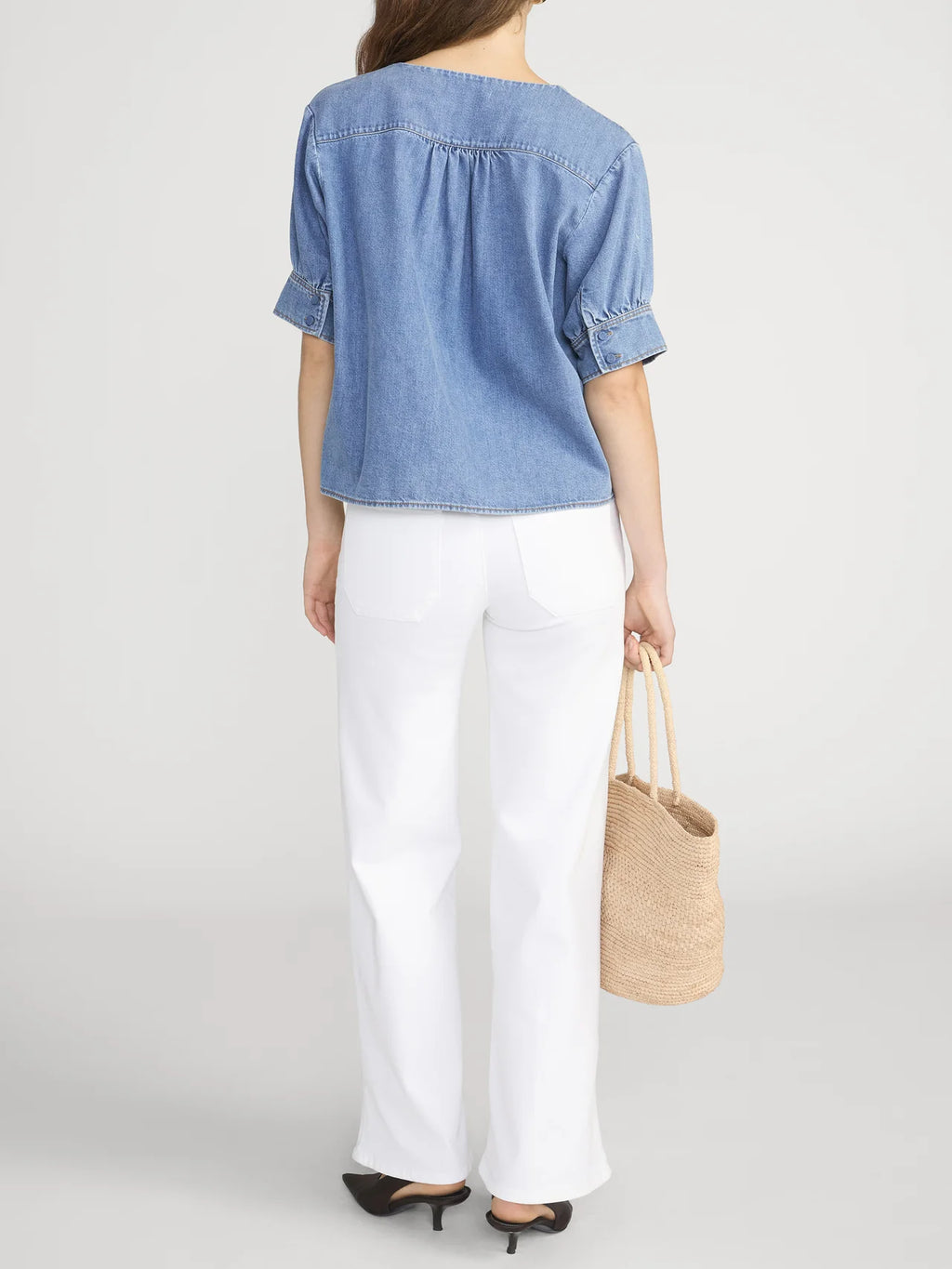 Woman wearing a blue denim shirt and white pants, holding a straw bag on a plain background