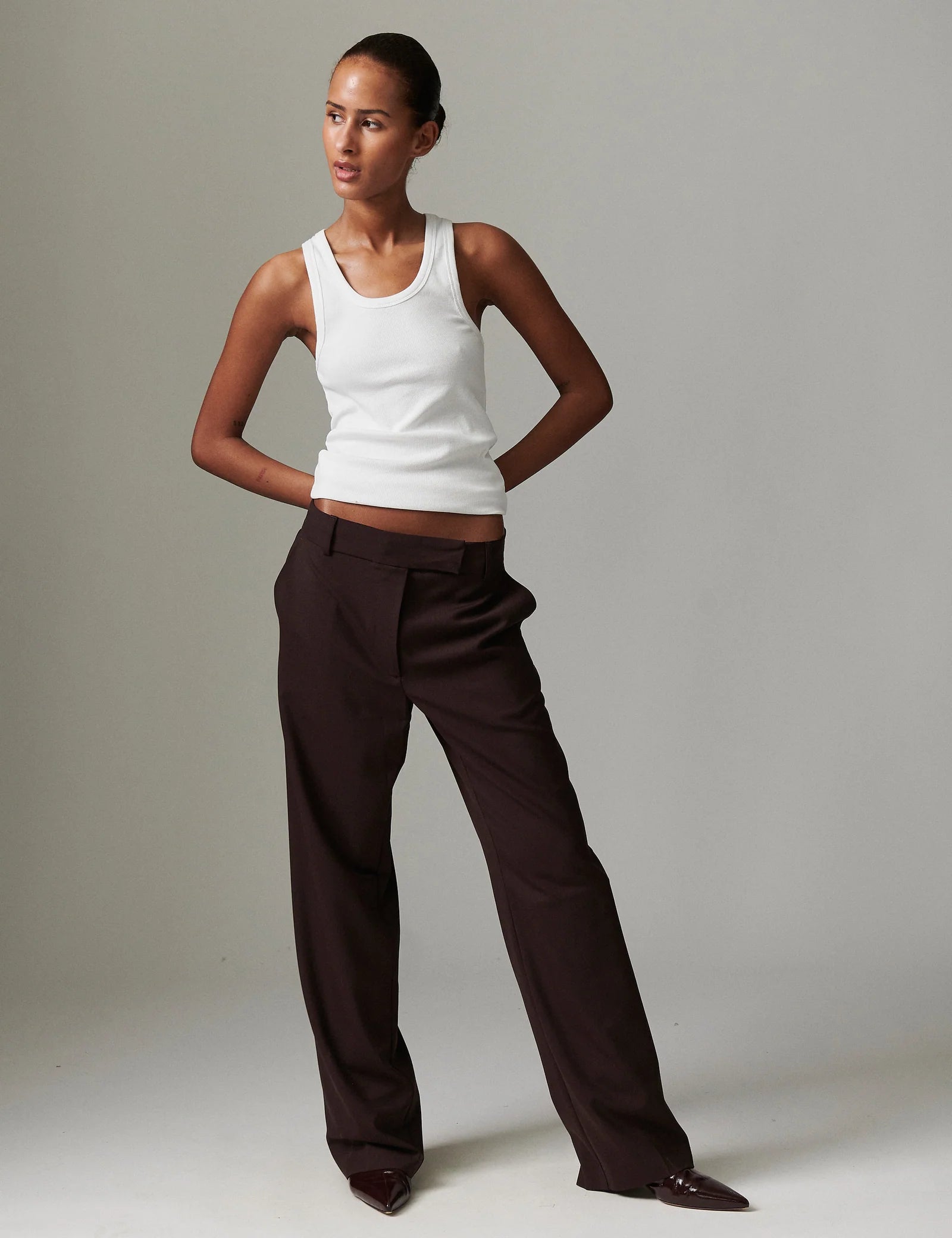 Woman wearing a white tank top and brown pants against a plain background