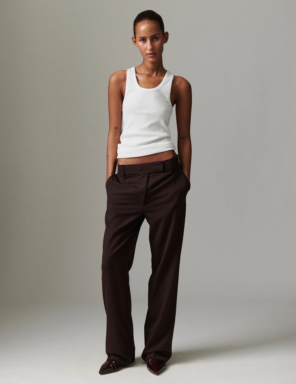 Woman wearing a white tank top and brown pants against a plain background