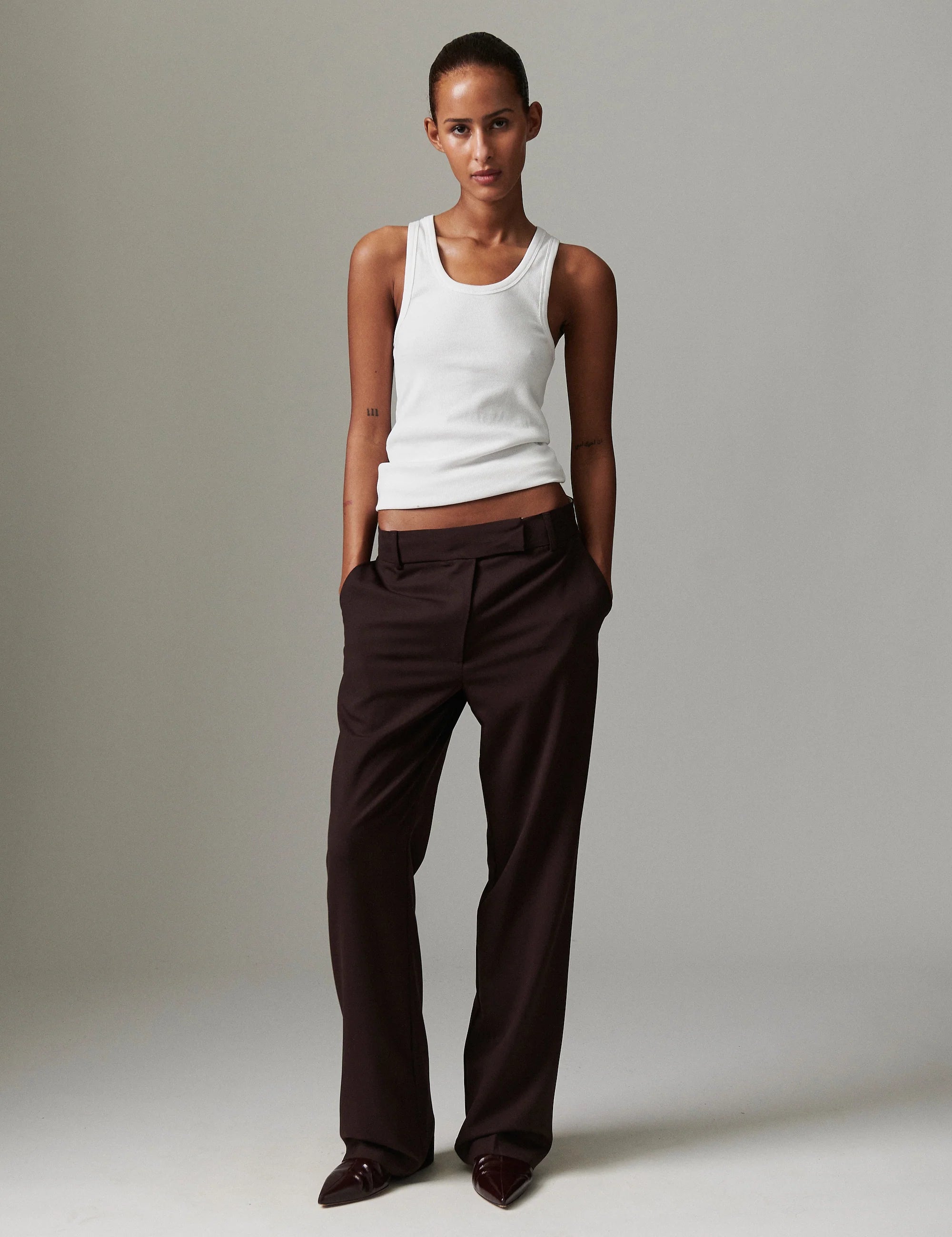 Woman wearing a white tank top and brown pants against a plain background