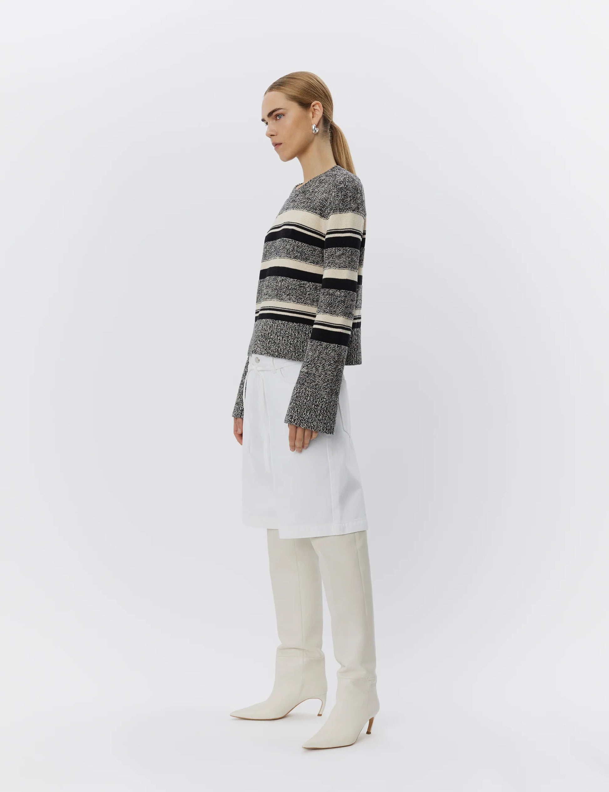 Woman wearing a striped sweater, white pants, and white boots on a plain background