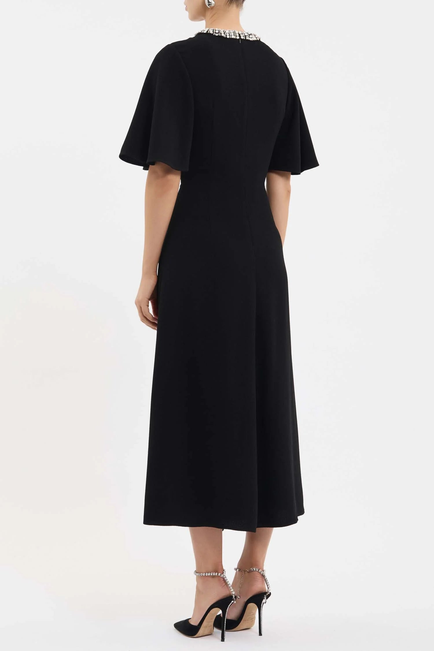 A woman shown from the back wearing a black midi dress with short flutter sleeves and a jeweled collar, paired with black high heels against a plain white studio background.