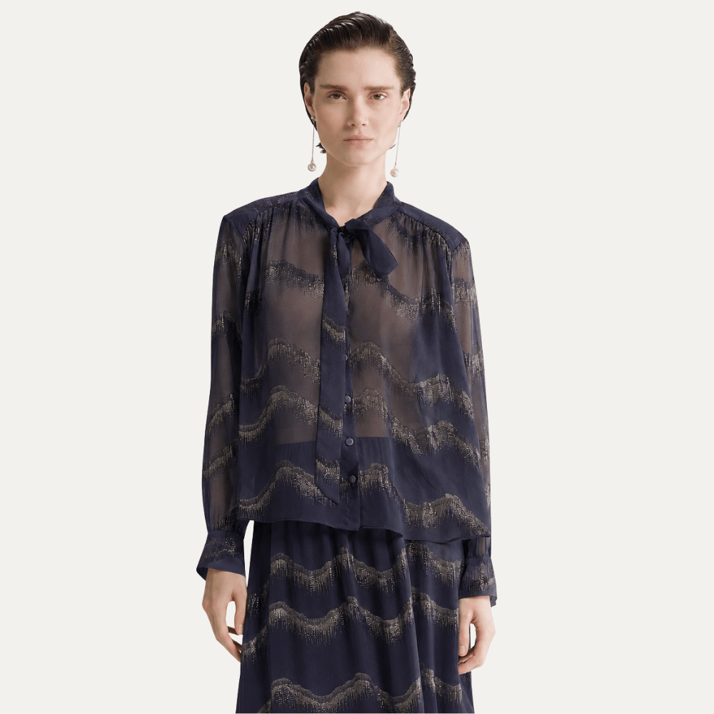A model wearing a sheer long-sleeve button-up blouse with a tie collar and a matching skirt, both featuring a dark navy blue and beige wavy abstract pattern, set against a plain light gray background.